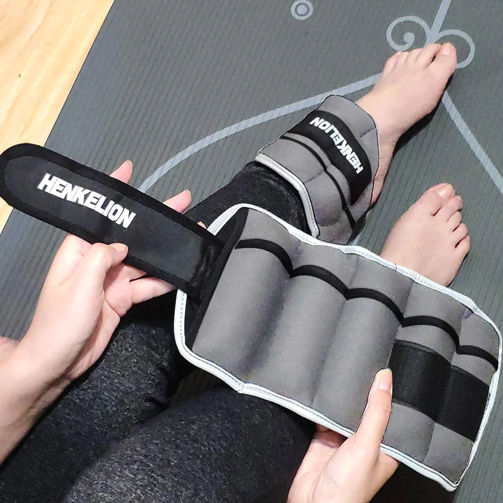 A gray Henkelion ankle weight opened flat on a yoga mat to show its row of weight pockets, with a second cuff already strapped around the other ankle.